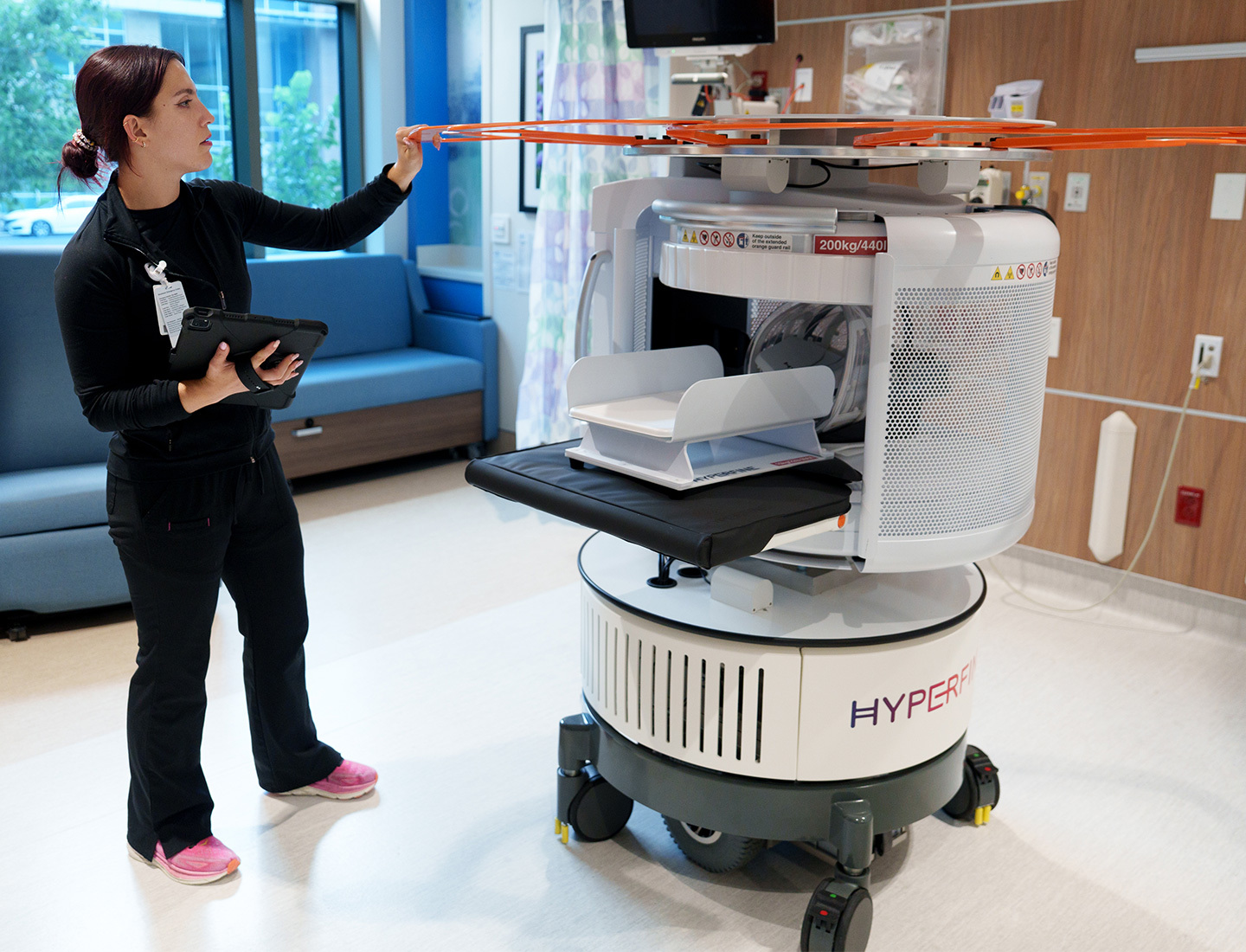 A woman wearing black jeans and a black long sleeve shirt is holding a tablet in her right hand, and her left hand is touching the top of a mobile MRI machine. The MRI machine is on wheels and is mostly white with a large orange circular structure at the top.
