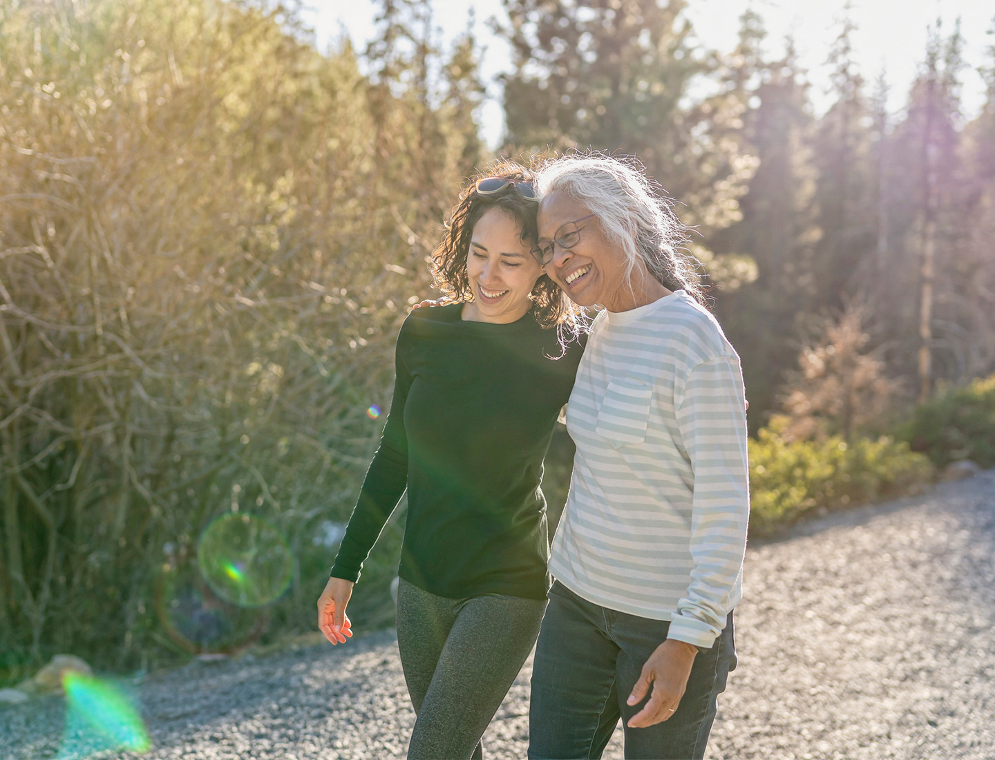 A woman and her retirement age mother walk arm in arm on a hiking trail surrounded by trees on a sunny day.