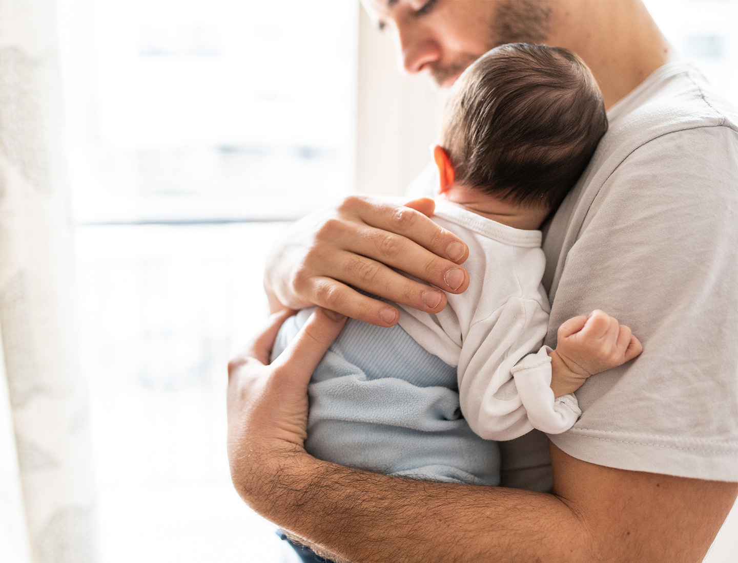 Adult male holding an infant close to his chest in a naturally lit room near a window.