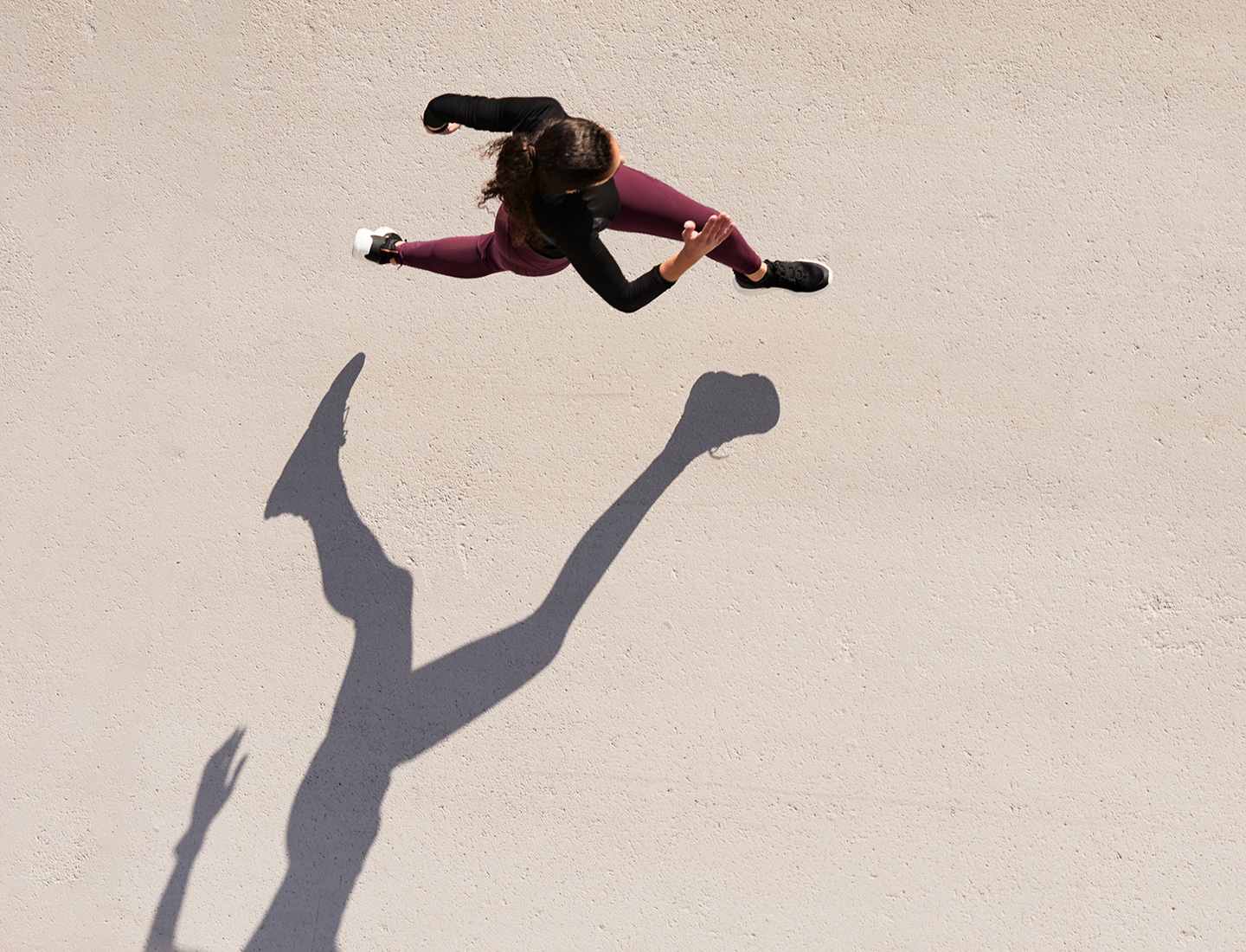 A young woman is seen sprinting from above in the bright sun.  She has dark brown hair, and is wearing a black top with purple leggings.  The sun casts a long shadow of her running.