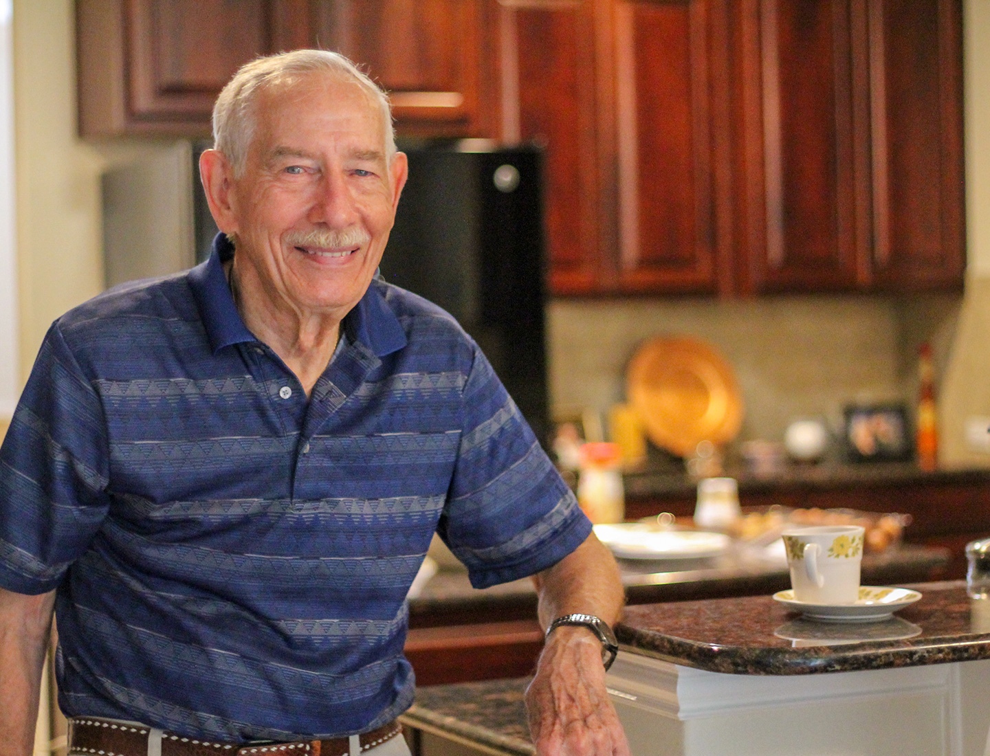 A senior man with a mustache stands in a kitchen, his arm resting on a countertop. He is smiling at the camera and wears a blue striped polo shirt.