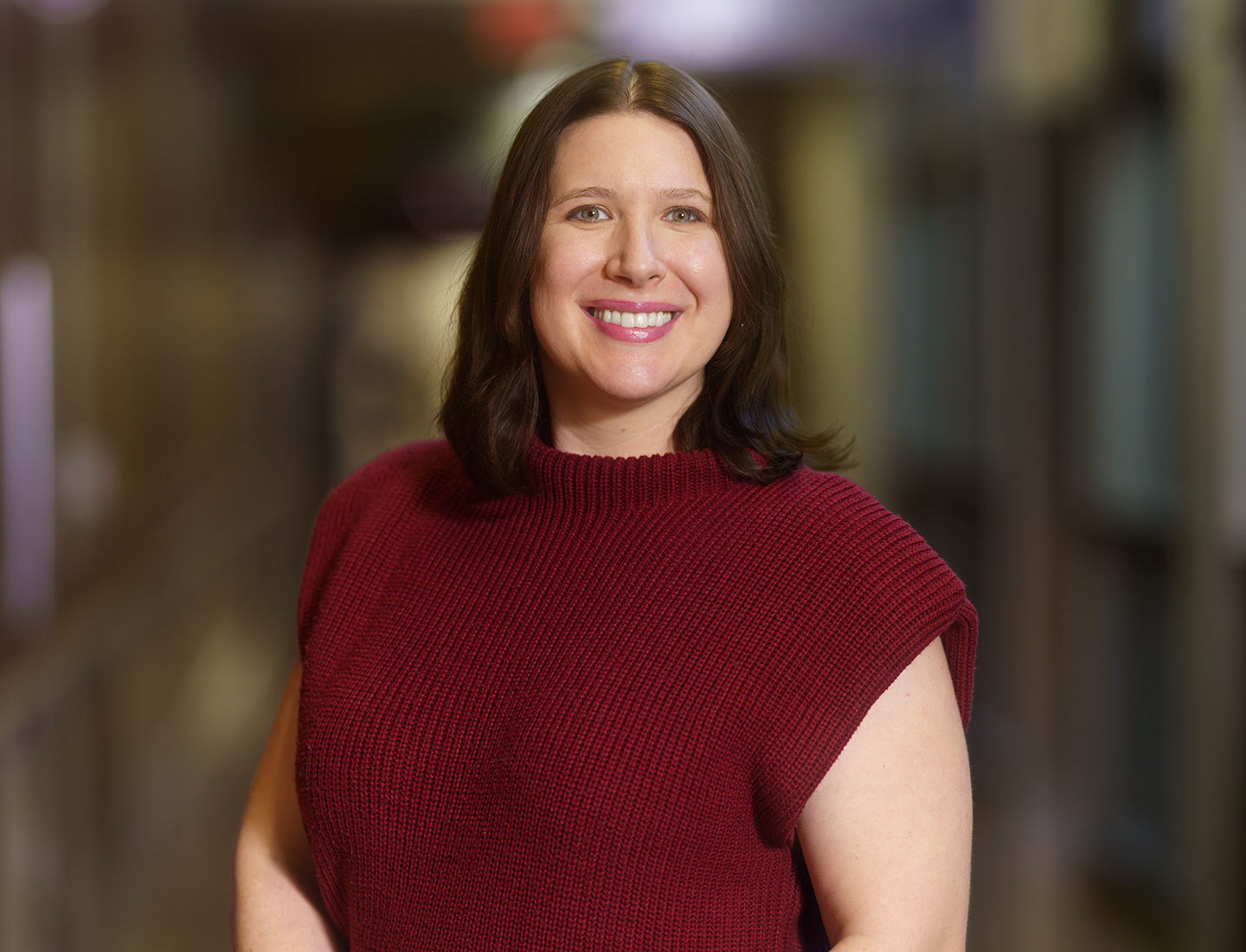 A closeup portrait of Chelsea Ortman. She is standing in the middle of a hallway and smiling at the camera. She is wearing a red sweater. image