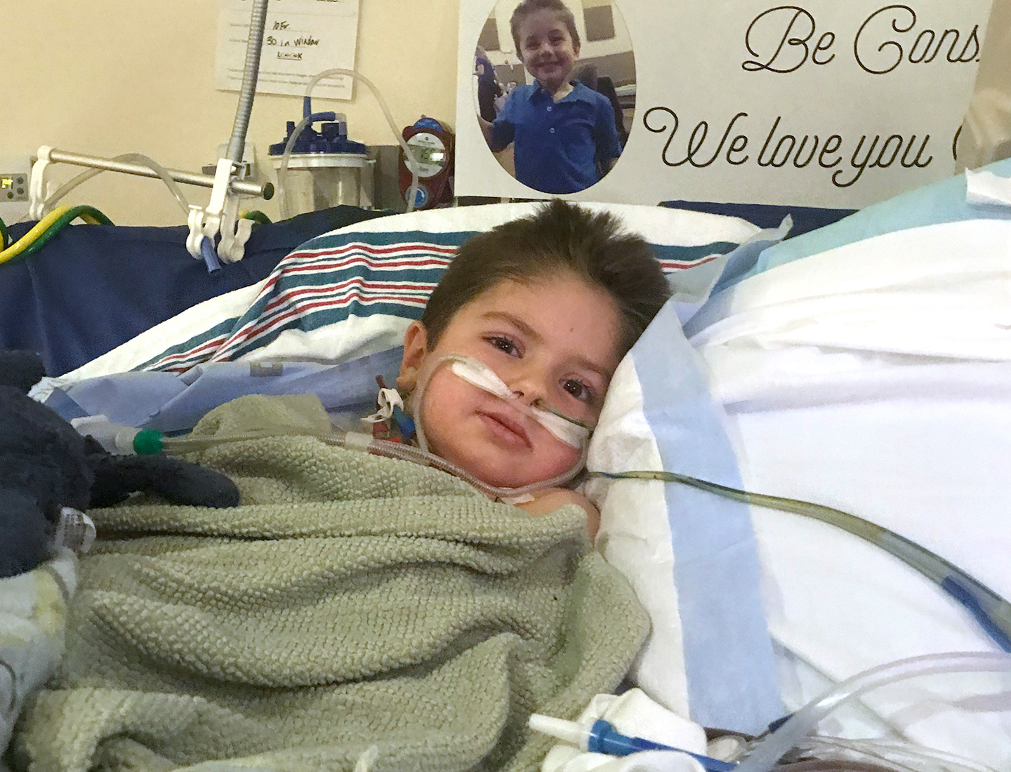 Owen Coulter lies in his hospital bed wrapped in an olive green blanket with a sign behind him that reads, "We love you".