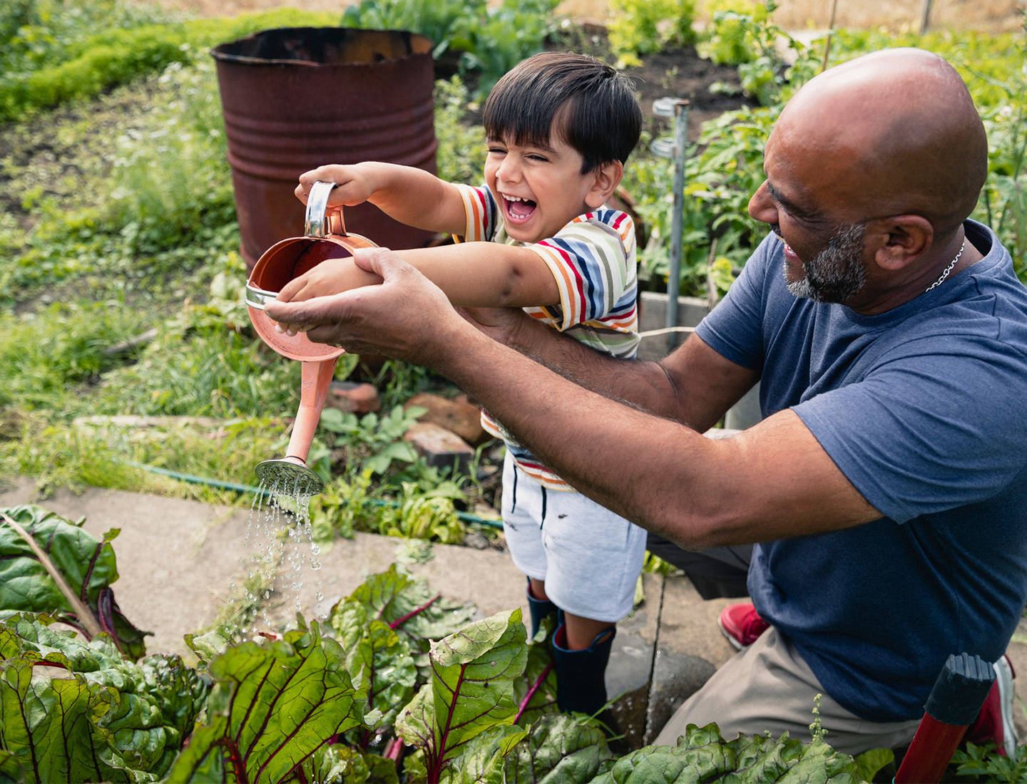 A man kneeling to help his son use a watering can on leafy plants.