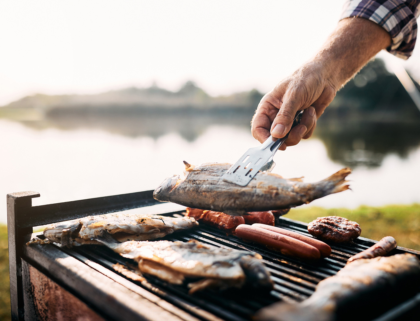 A closeup of whole fish on the grill. Someone is flipping one of the fish over with tongs, and a blurry lake is seen in the distance.
