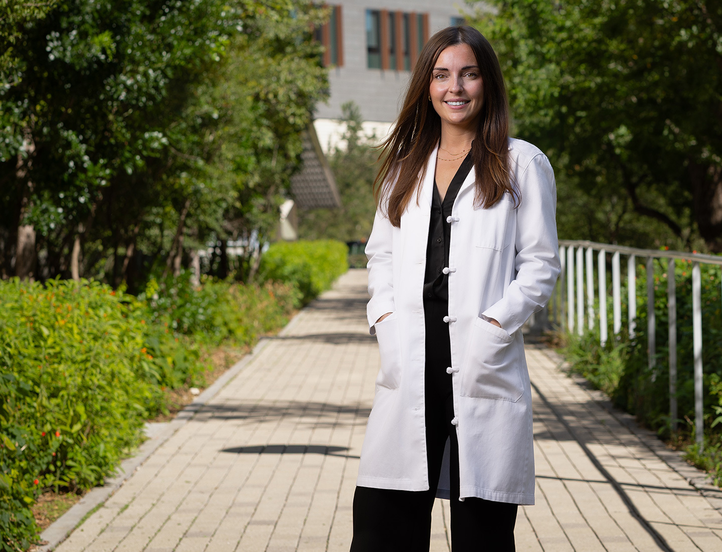 Dr. Elisa Furay is standing outside of the HLB buliding smiling at the camera. She is wearing a black blouse and pants and a white coat. image