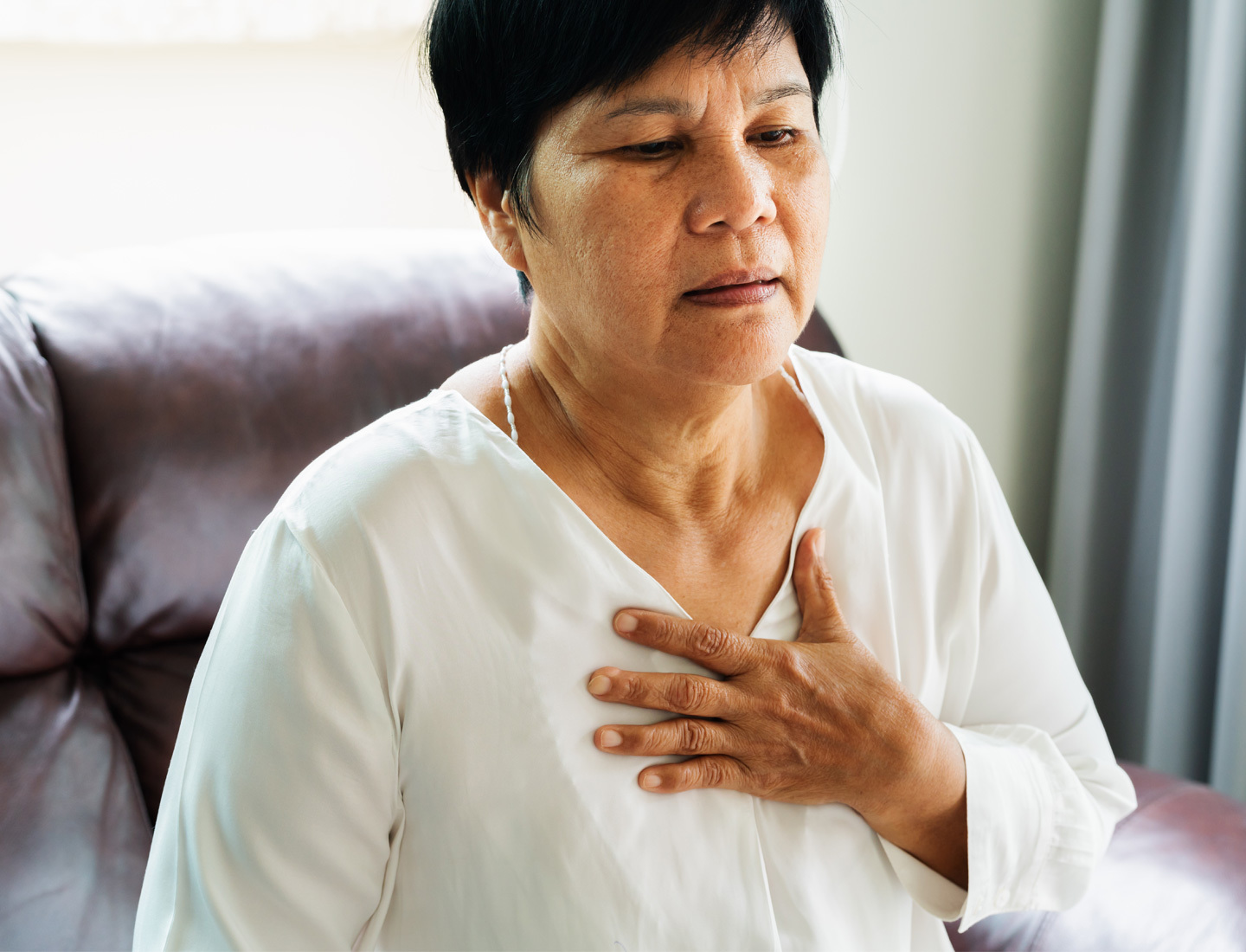 A seated Latin-X woman experiencing heartburn holds her hand to her chest.