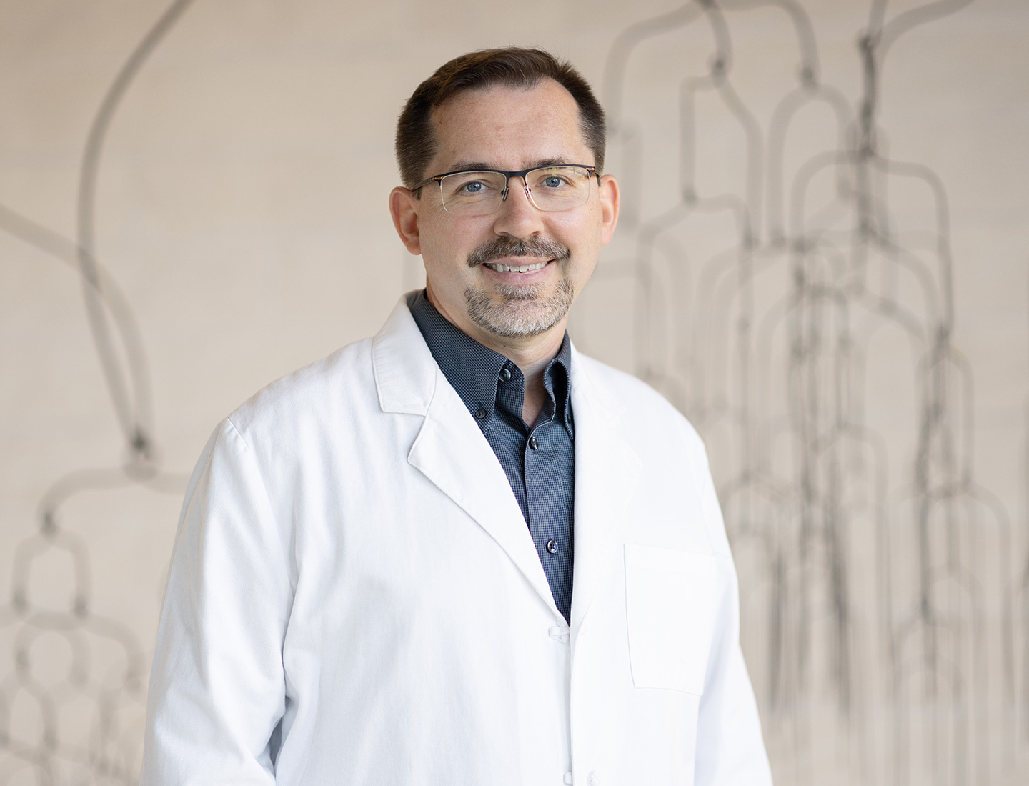 Dr. Jon Willie is smiling at the camera and standing in front of a large wire-like metal sculpture. He is wearing a navy dress shirt and a white coat. image