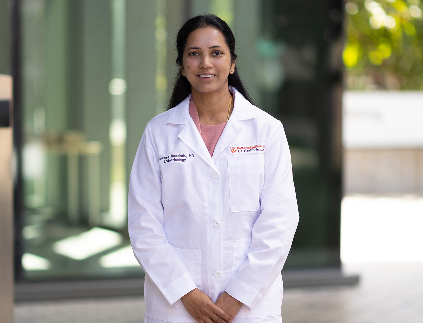 Dr. Sowmya Boddhula is smiling at the camera and wearing a pink dress suit with a white coat over it. She is standing outside in the courtyard between the HTB and HDB buildings.