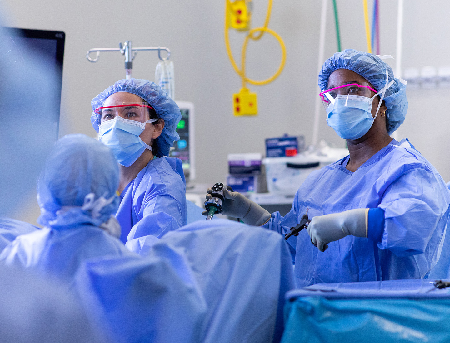 A group of clinicians wear surgical gowns, masks, hairnets, and protective eyewear in the operating room.