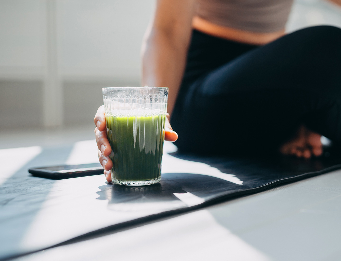 A close up of a glass of a green smoothie, being held by a woman seated on a yoga mat.