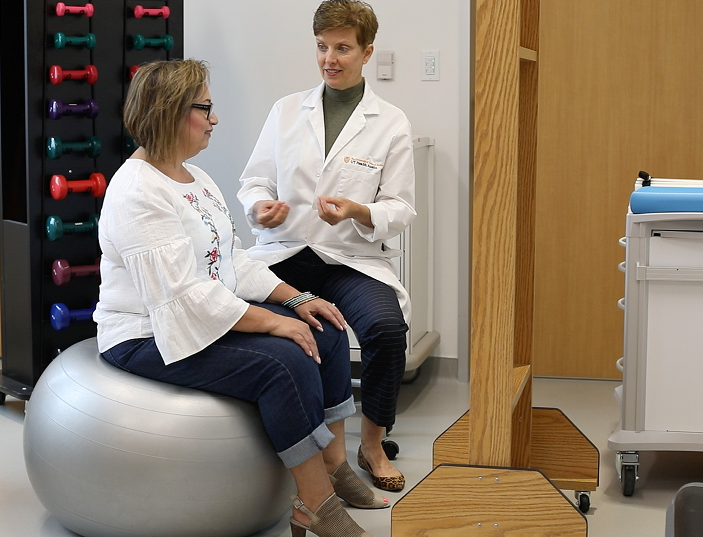 A seated UT Health Austin clinician speaking to a woman who sits on a Swiss ball.