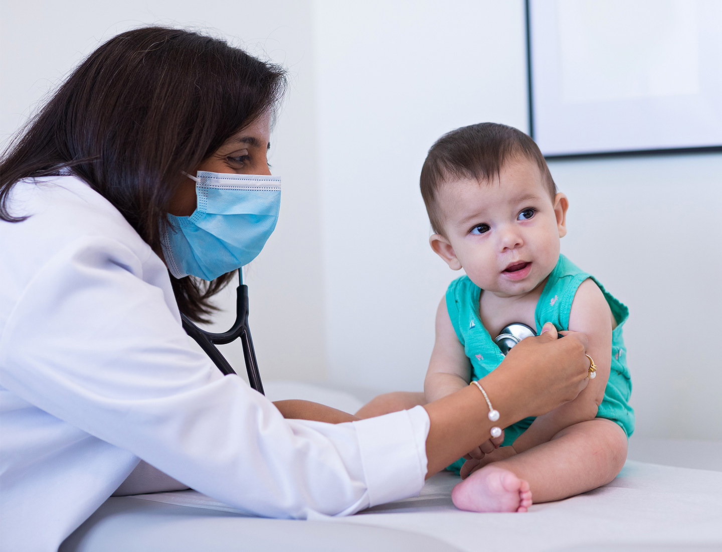A female doctor in a white coat wearing a surgical mask listens to an infant's heartbeat using a stethoscope.