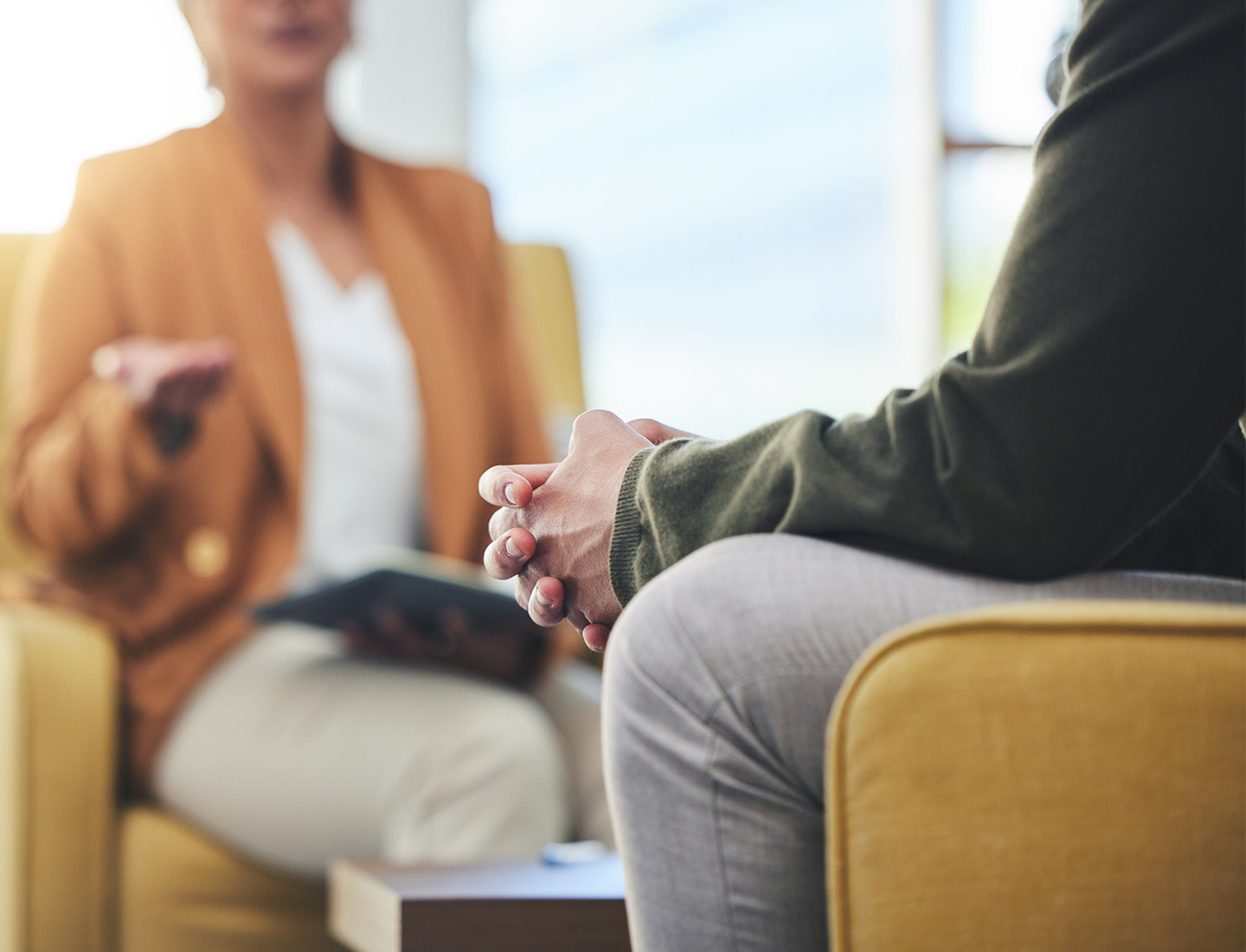 Two people sit across from each other in conversation, one with hands clasped, the other gesturing.