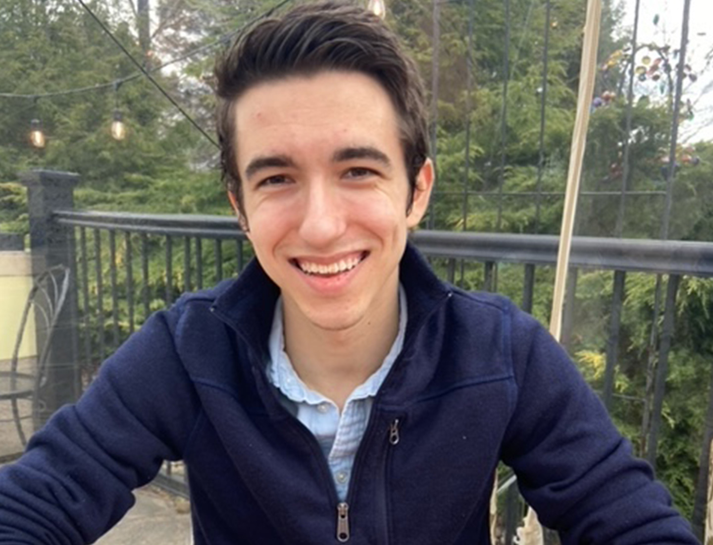 Dom Rainelli is wearing a blue jacket, looking at the camera, and smiling. He is sitting outside on a deck on an overcast day.