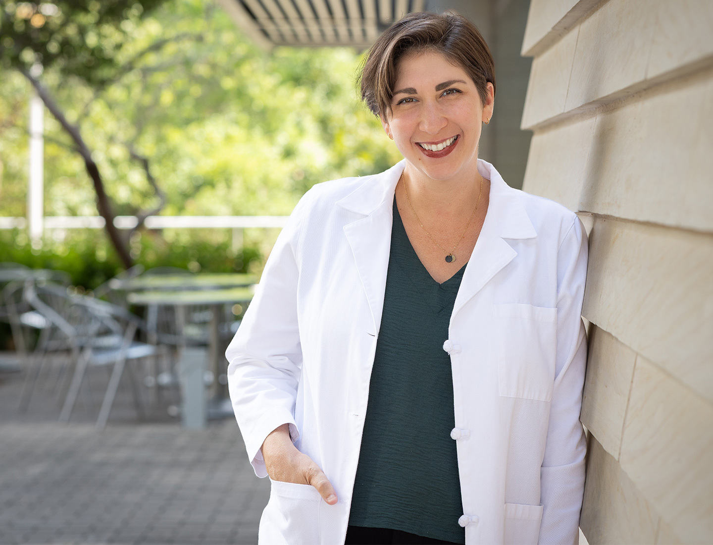 An outdoor portrait of Dr. Elizabeth Rosenthal. She is smiling and leaning against a building wall, and has one hand in her white coat pocket. The setting is in a courtyard on a bright, sunny day. image