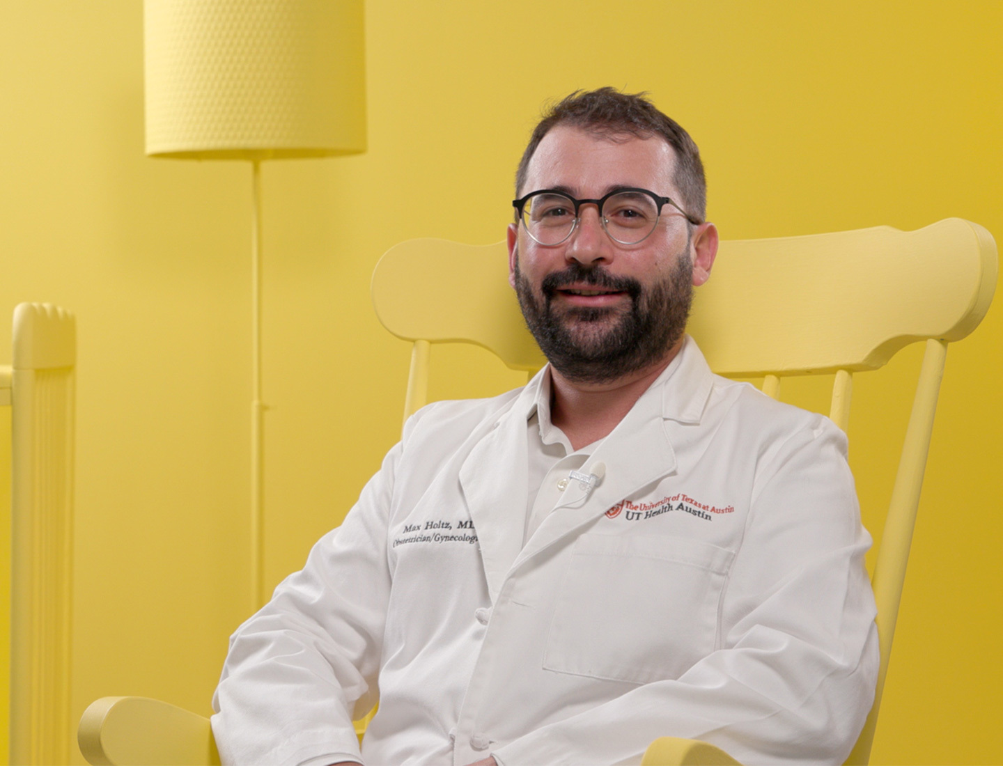 A physician wearing a white coat and glasses sits in a bright yellow rocking chair in a yellow nursery.