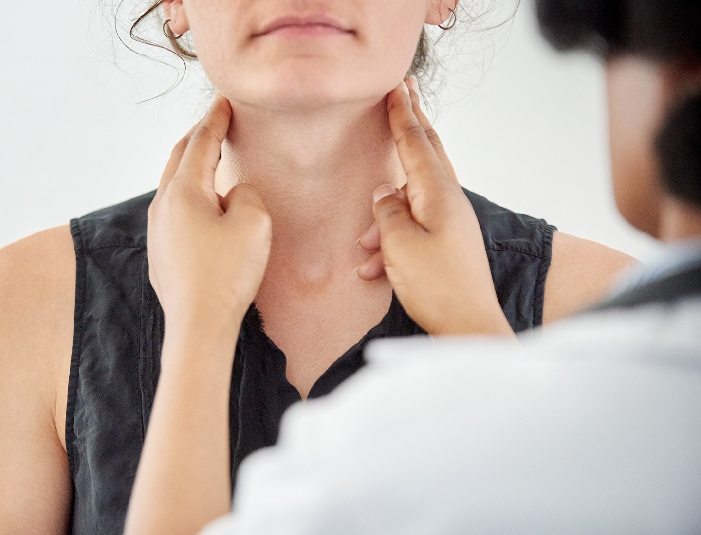 A closeup is shown of a female patient in a doctors office. The doctor's hands are on either side of the patient's neck just under her jaw line for a thyroid check up.