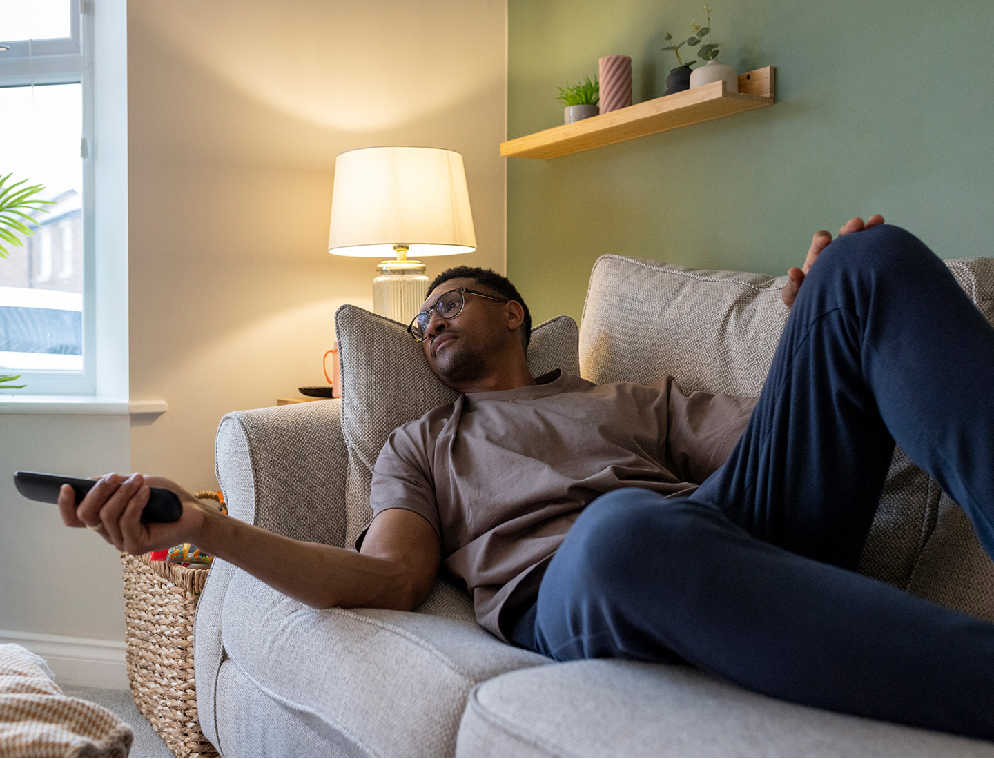 Adult man reclines on a couch holding a remote control, resting at home in a calm, softly lit room.