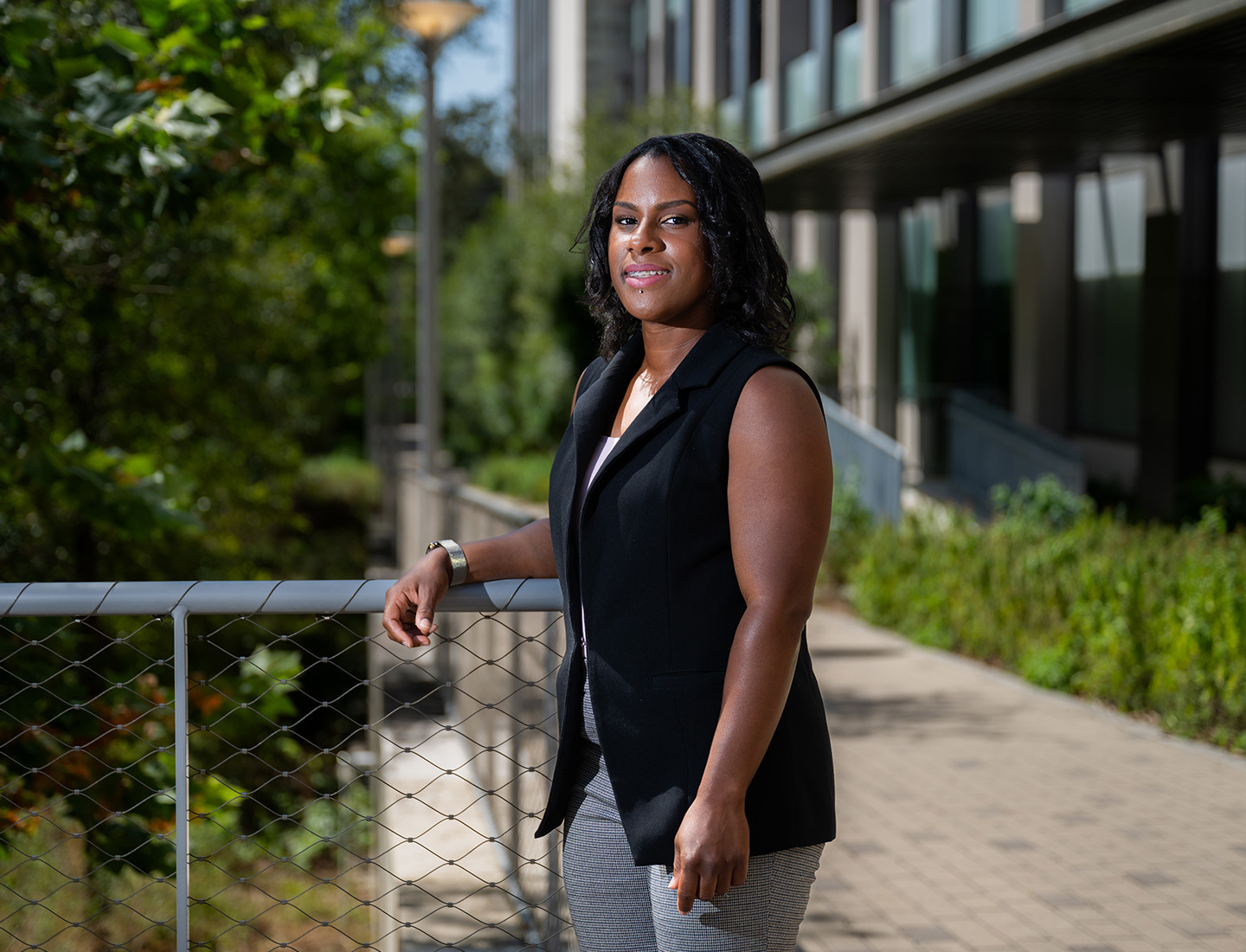 Deon Davis is standing outside smiling at the camera and resting her arm on a fence.  She is wearing a black vest and grey pants. image