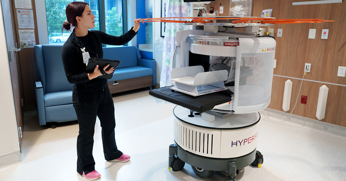 A woman wearing black jeans and a black long sleeve shirt is holding a tablet in her right hand, and her left hand is touching the top of a mobile MRI machine. The MRI machine is on wheels and is mostly white with a large orange circular structure at the top.