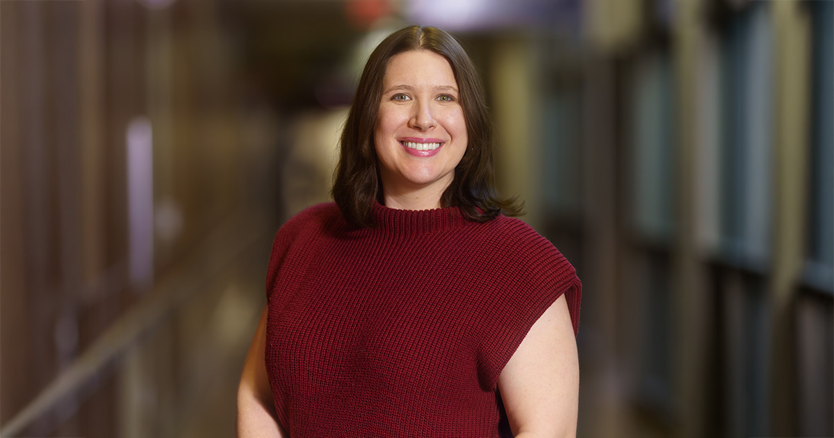 A closeup portrait of Chelsea Ortman. She is standing in the middle of a hallway and smiling at the camera. She is wearing a red sweater.