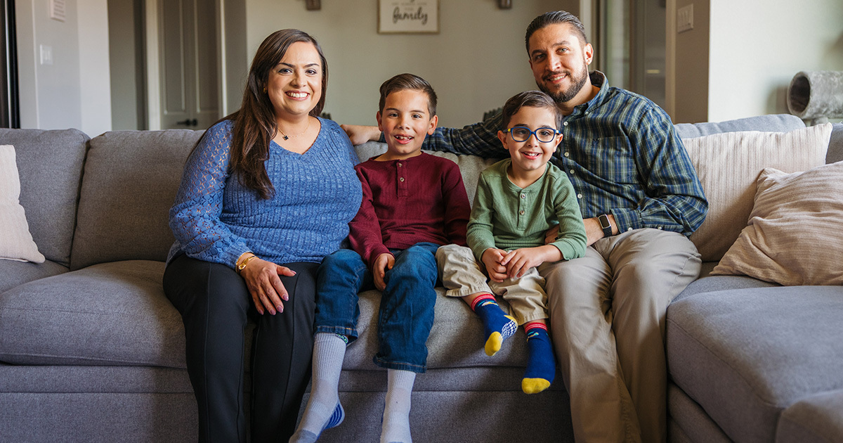 The Arouh family is sitting on their gray couch in their living room. The mom and dad are seated on either side of their two little boys. They are smiling at the camera.