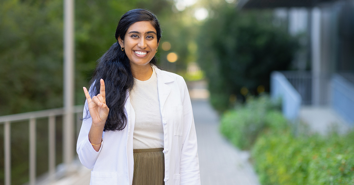 Dr. Divya Rayapati is smiling at the camera and holding her right hand up in the hook 'em formation. She is standing outside of the HLB and is wearing a cream colored sweater, olive green skirt, and a white coat.