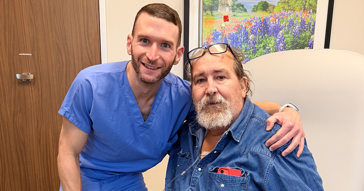 A photo of Dr. Grimm and Ray smiling for the camera. Dr. Grim has his arm around Ray, who is seated in a doctor's office. Dr. Grimm is wearing blue scrubs, Ray is wearing blue denim, and a bluebonnet painting is behind them.