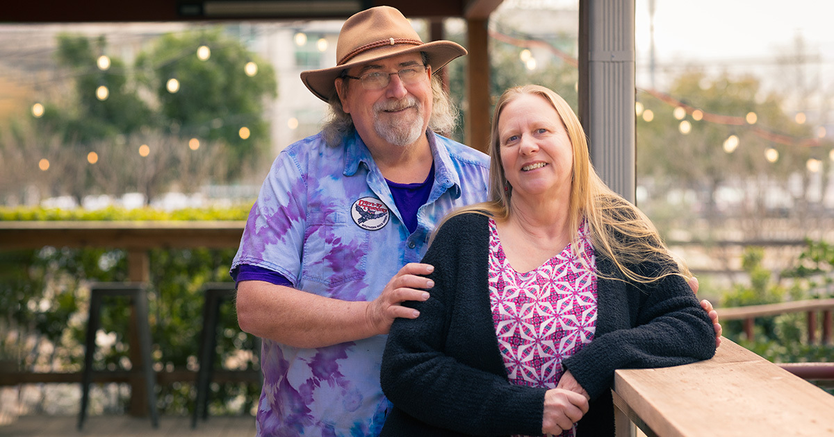 Trey and Mickie Bailey are standing outside on a porch and smiling at the camera. Trey is wearing a blue and purple tie die shirt and a hat, and Mickie is wearing a purple shirt with a black cardignan.