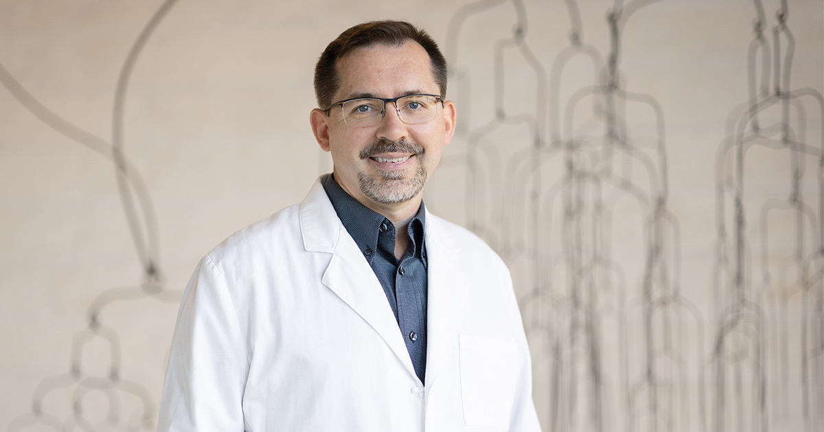 Dr. Jon Willie is smiling at the camera and standing in front of a large wire-like metal sculpture. He is wearing a navy dress shirt and a white coat.
