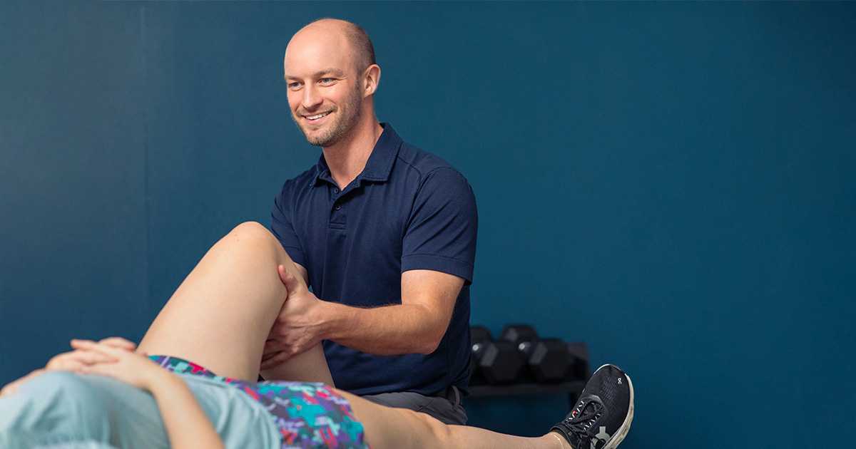 Smiling physical therapist supports a patient’s bent leg during a rehab session in a clinical setting.