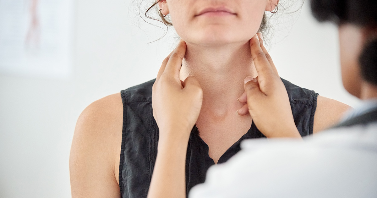 A closeup is shown of a female patient in a doctors office. The doctor's hands are on either side of the patient's neck just under her jaw line for a thyroid check up.