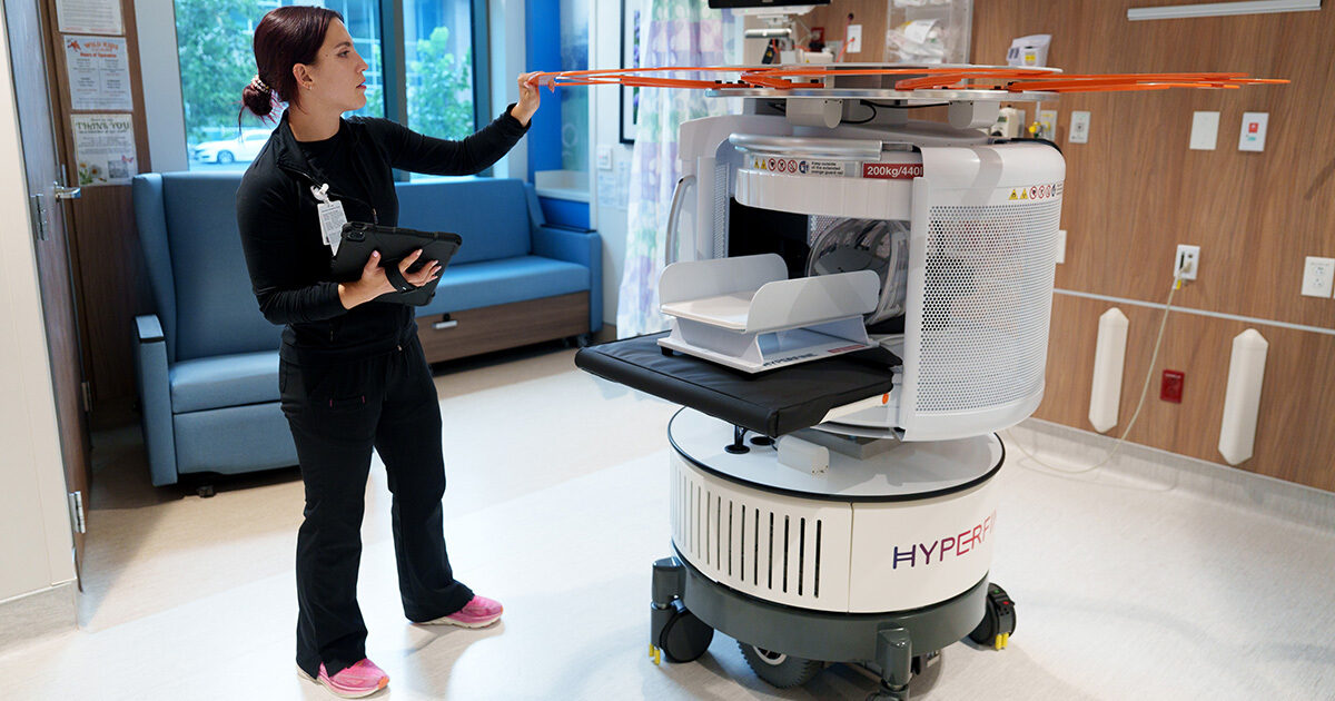 A woman wearing black jeans and a black long sleeve shirt is holding a tablet in her right hand, and her left hand is touching the top of a mobile MRI machine. The MRI machine is on wheels and is mostly white with a large orange circular structure at the top.
