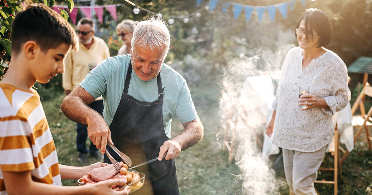 An older man wearing a black apron and a green shirt hands a younger kid a plate of barbecued food. Another middle aged woman looks on and smiles. They're standing around a grill at an outdoor backyard party.