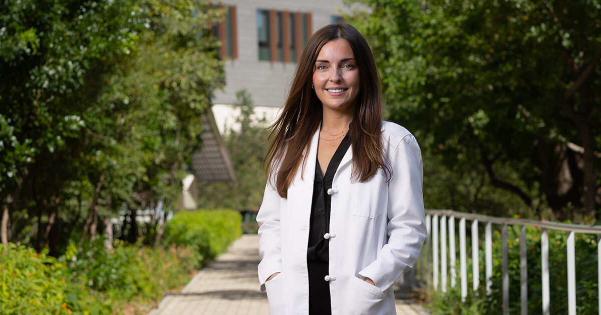 Dr. Elisa Furay is standing outside of the HLB buliding smiling at the camera. She is wearing a black blouse and pants and a white coat.