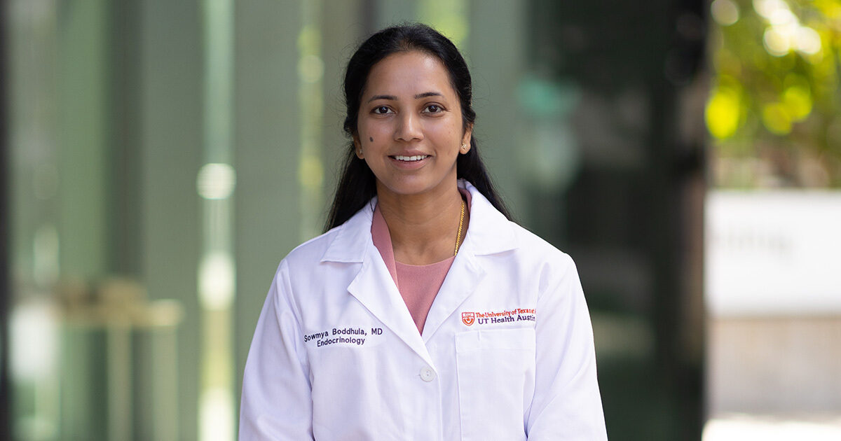 Dr. Sowmya Boddhula is smiling at the camera and wearing a pink dress suit with a white coat over it. She is standing outside in the courtyard between the HTB and HDB buildings.