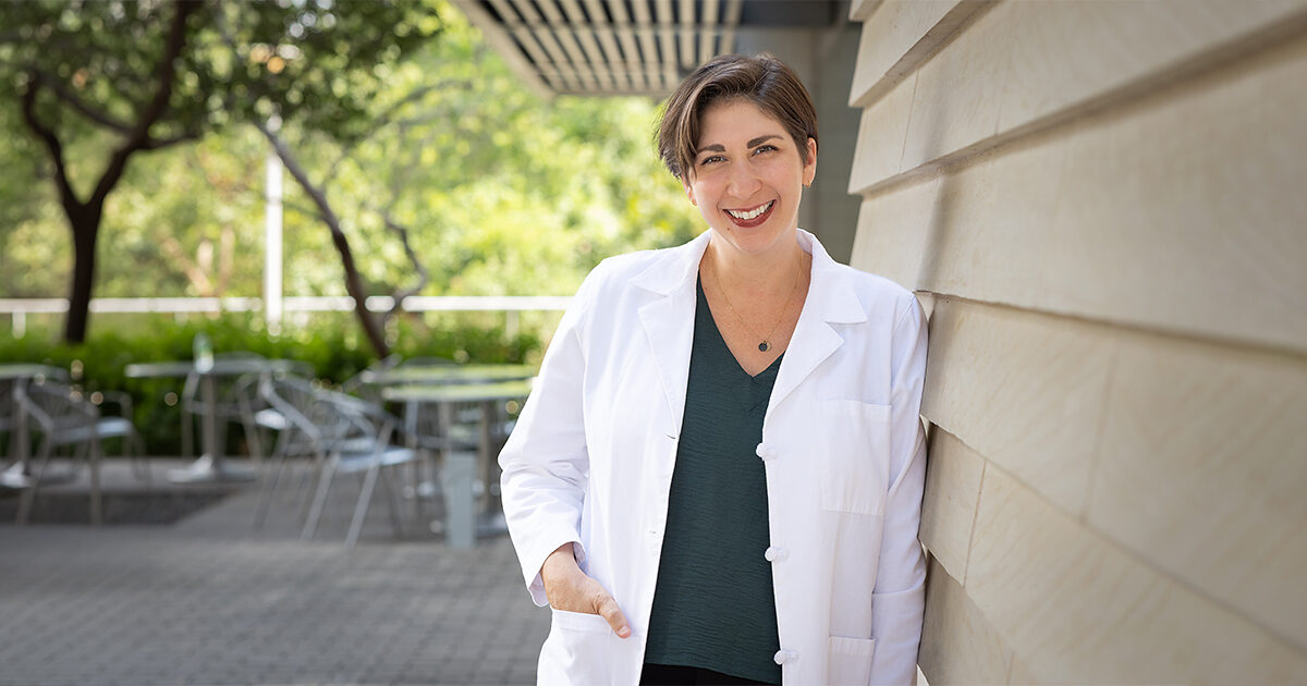 An outdoor portrait of Dr. Elizabeth Rosenthal. She is smiling and leaning against a building wall, and has one hand in her white coat pocket. The setting is in a courtyard on a bright, sunny day.