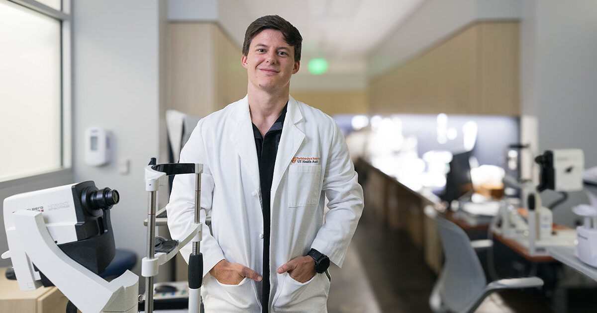 Dr. Kirby Taylor is wearing a white coat and has his hands in his pockets. He is standing behind ophthalmology equipment in an imaging lab.