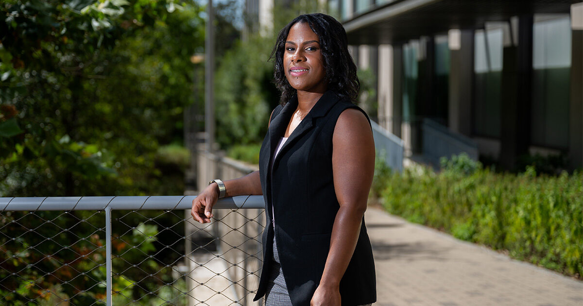 Deon Davis is standing outside smiling at the camera and resting her arm on a fence.  She is wearing a black vest and grey pants.