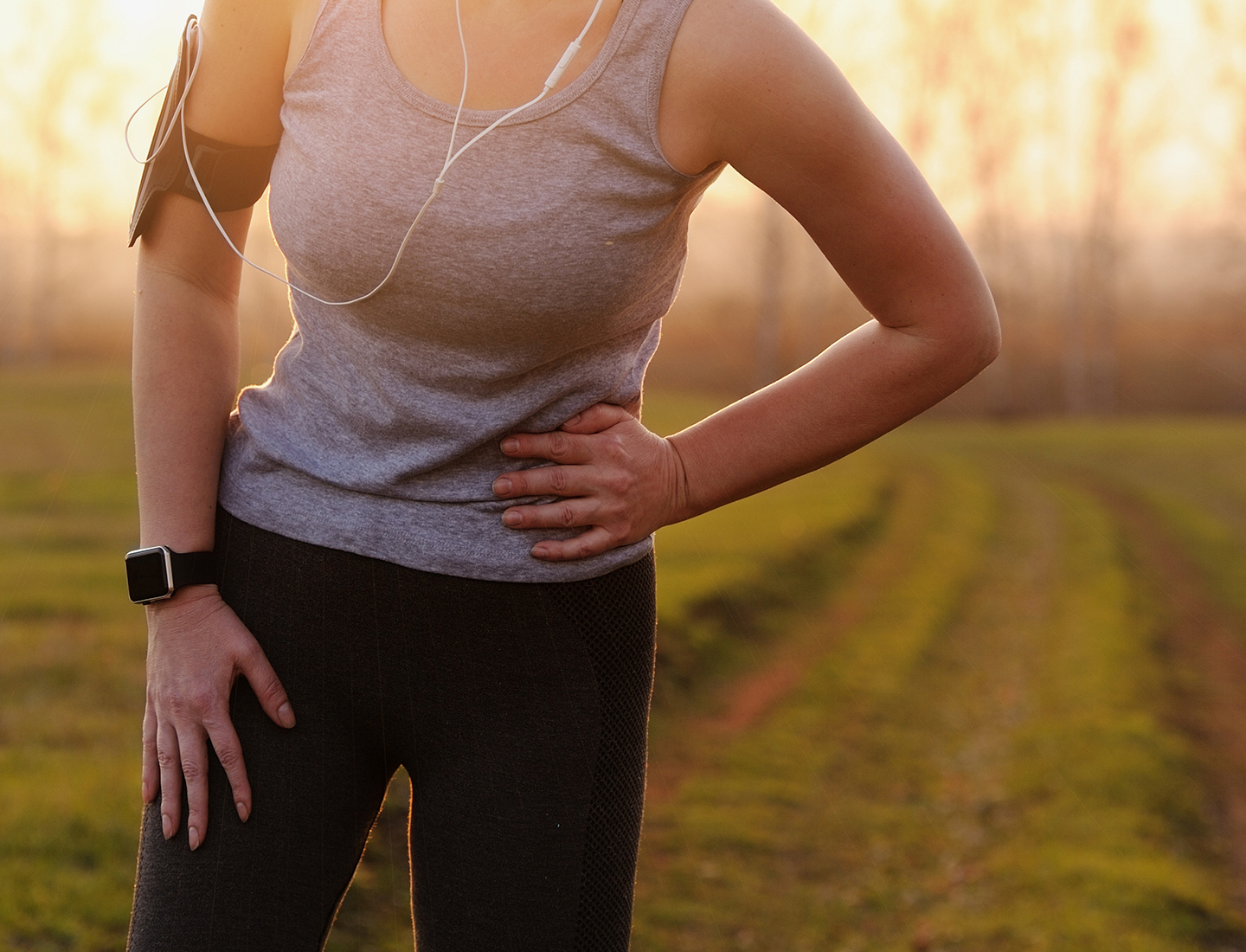 A closeup view of a woman wearing athletic clothes, standing outside, while she holds the side of her abdomen in an expression of discomfort.