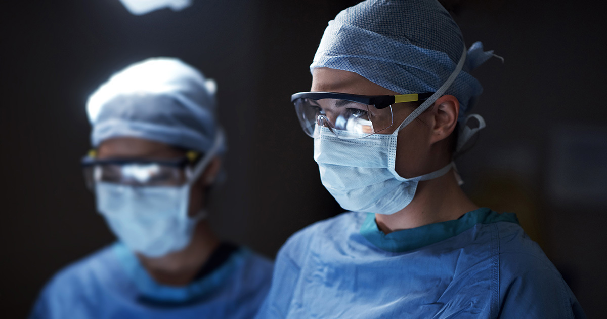 Two surgeons wearing masks, caps, and protective glasses focus during an abdominal procedure.