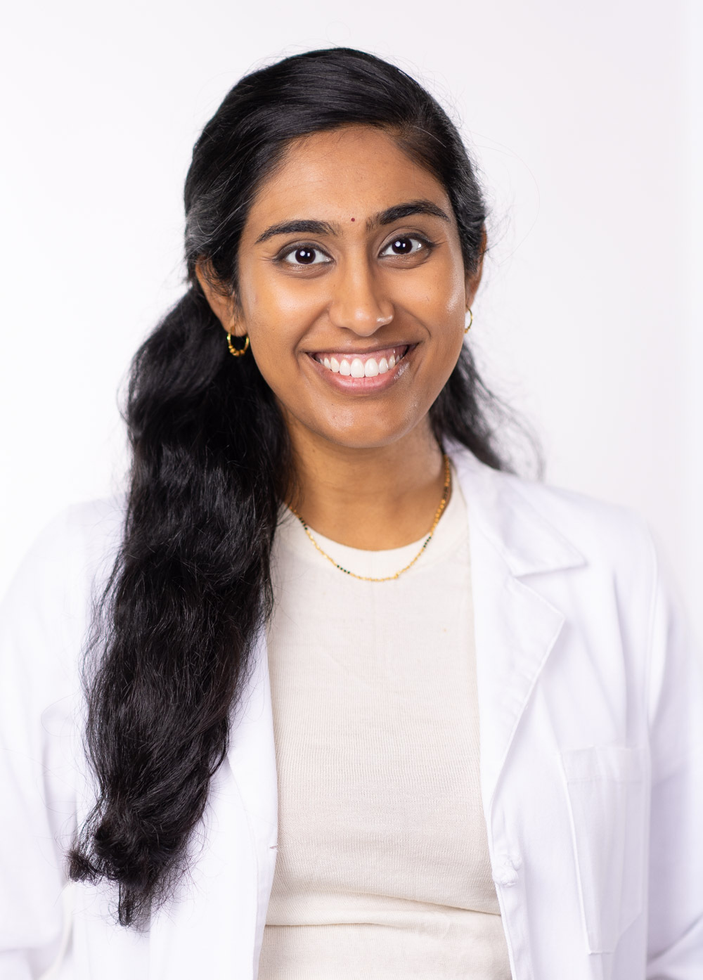 Dr. Divya Rayapati is smiling at the camera. She is wearing a beige sweater and a white coat.