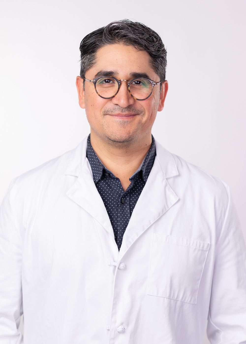 Headshot of Joaquin Camara. He is wearing a white coat and a dark navy dress shirt with small white polka dots.