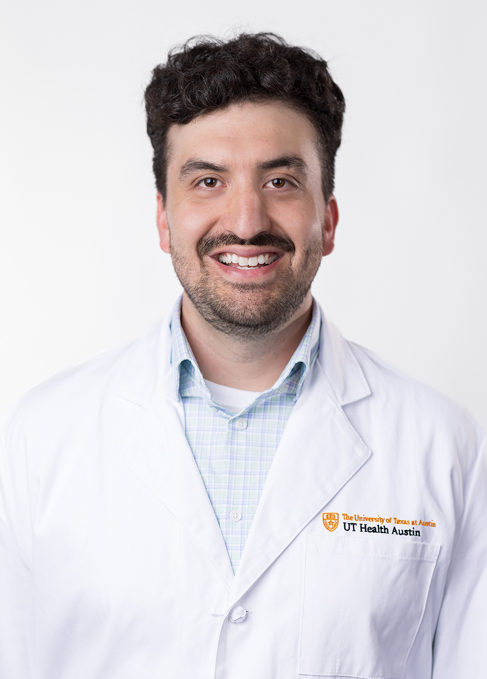 Dr. Jordan Jacquez is smiling at the camera and wearing a white coat with a blue plaid dress shirt.
