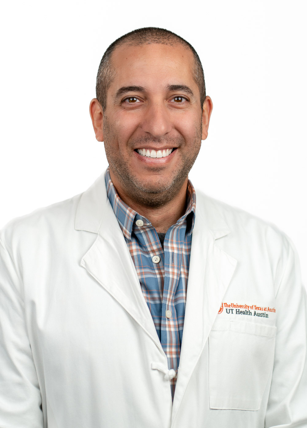 Justin Solomon is smiling at the camera. He is wearing a white coat and a blue and red plaid dress shirt.